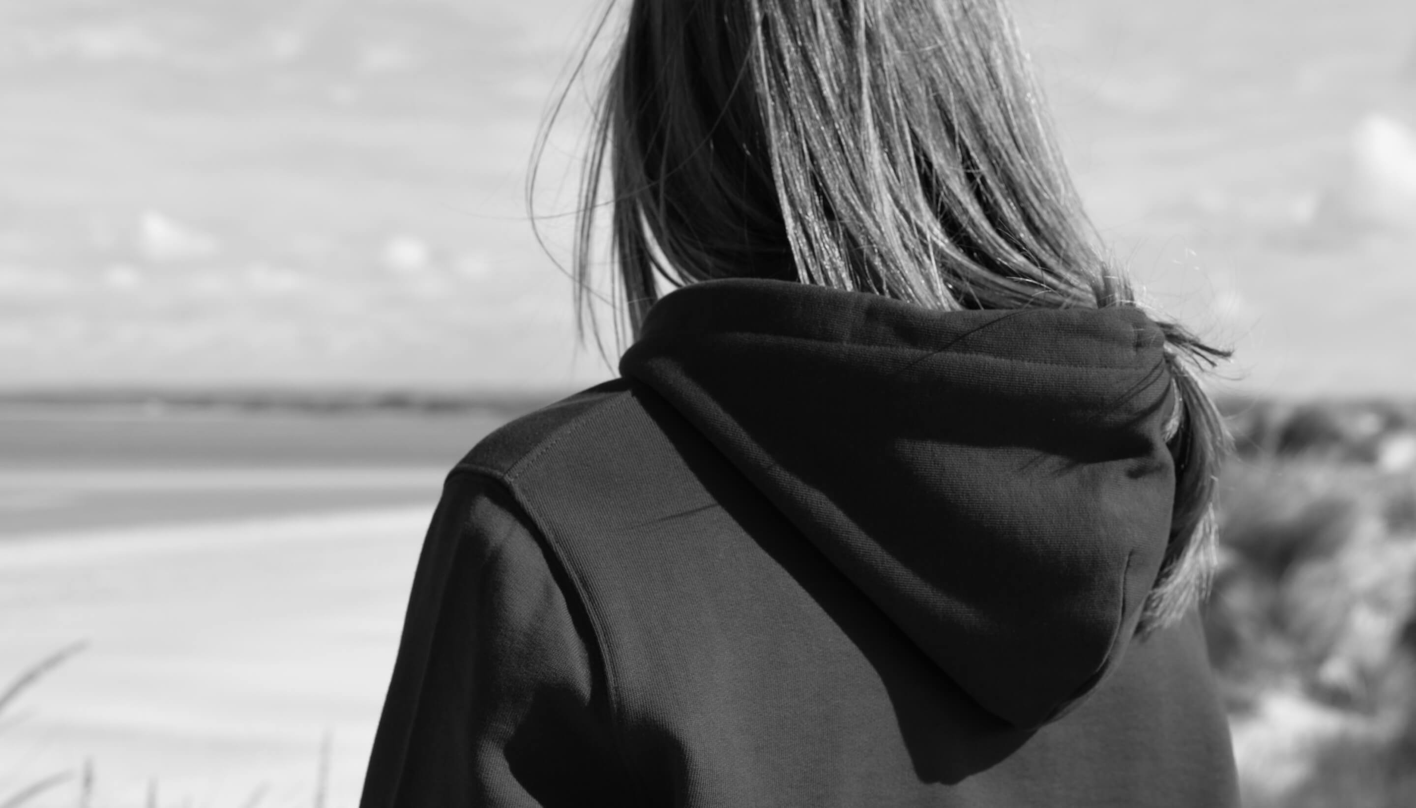 Closeup of a women viewed from the back wearing a hoodie with a beach in the background. The image is in black and white.